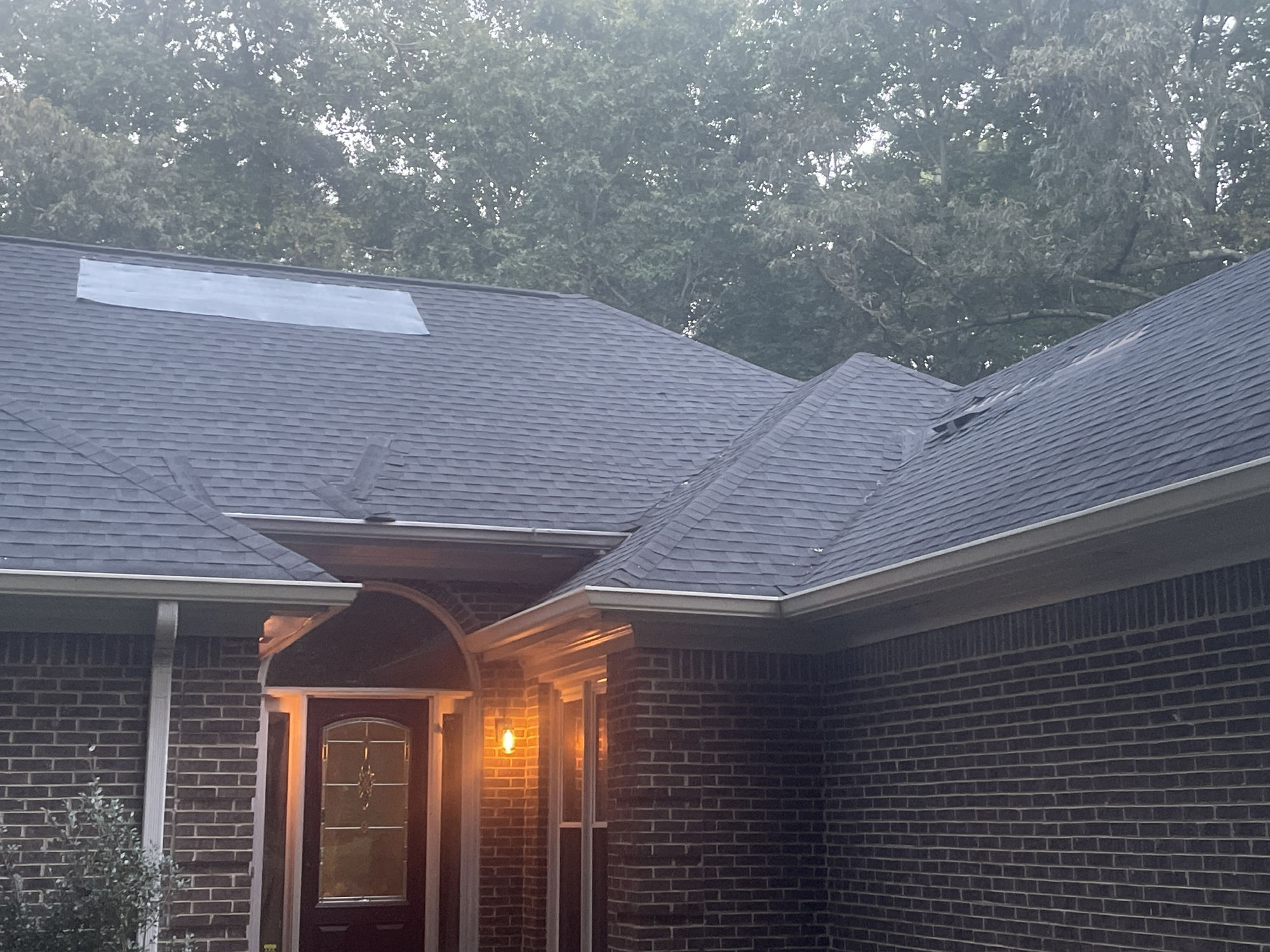 Front entry at dusk. Patch section on the upper left slope and torn, displaced shingles on the right slope above the gutter.