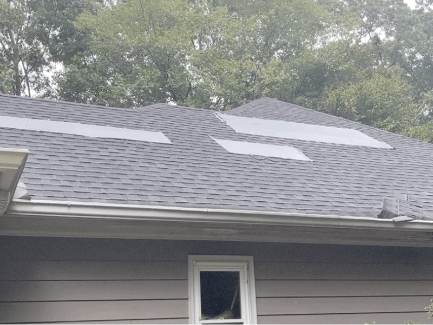 Rear slope, wide view. Multiple gray patch areas where failed shingle sections were covered. Damaged shingle debris visible at the right eave over the gutter.